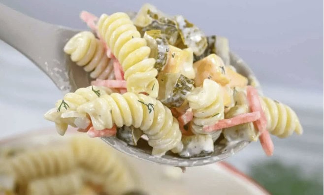 Close-up of a spoonful of rotini pasta salad with diced vegetables, pickles, and creamy dressing—a perfect side for backyard barbecue gatherings.